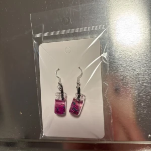Rosa örhängen med glittrig design - Snygga örhängen i rosa med en glittrig design. De har en unik form som liknar små flaskor fyllda med rosa vätska och glitter. Perfekta för att ge en färgklick till din outfit. (Billigt material) 