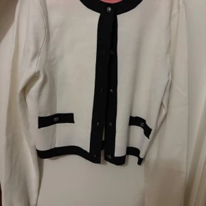 Vit kofta med svarta detaljer - Säljer en stilren vit kofta med svarta kanter och knappar. Koftan har långa ärmar och två dekorativa fickor framtill. Perfekt för en elegant look.