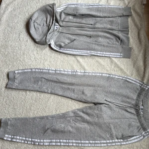 Grå mjukisdress från Adidas - Säljer ett par grå mjukisbyxor från Adidas med de klassiska tre ränderna längs sidorna. Byxorna har en elastisk midja med dragsko och muddar vid bensluten för en bekväm passform. Perfekta för en avslappnad stil. Säljer Dressen eller plaggen separat (100kr st eller 150kr för båda)