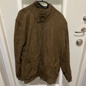 Brun mockajacka med dragkedja - Snygg brun mockajacka med dragkedja och hög krage. Jackan har långa ärmar och praktiska fickor med knappar framtill. Perfekt för höst och vår.