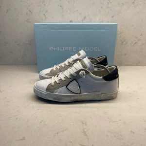 Vita sneakers från Philippe Model - Helt nya och oanvända vita sneakers från Philippe Model med grå och mörkblåa detaljer. Skorna har en klassisk design med snörning och en bekväm sula. Perfekta för en stilren look.