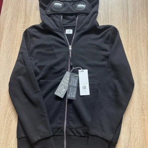 Svart huvtröja med glasögon från C.P. Company - Säljer en svart huvtröja från C.P. Company med unika glasögon i huvan. Tröjan har dragkedja framtill och ribbade muddar vid ärmslut och nederkant. Perfekt för en cool och avslappnad stil.