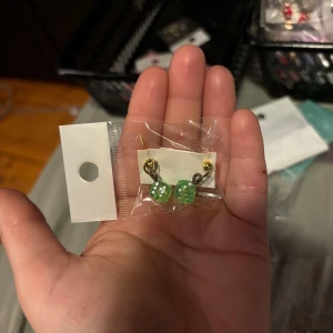 Gröna tärningsörhängen - Säljer ett par unika örhängen med gröna tärningar. De har en guldfärgad detalj och är förpackade i en plastpåse. Perfekta för att ge en lekfull touch till din stil.