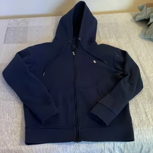 Mörkblå hoodie med dragkedja - Säljer en stilren mörkblå hoodie med dragkedja och justerbar huva. Tröjan har långa ärmar och en liten logga på bröstet. Perfekt för en avslappnad stil.