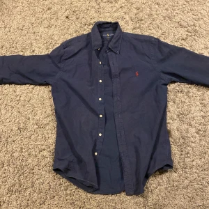 Mörkblå skjorta från Ralph Lauren - Säljer en stilren mörkblå skjorta från Ralph Lauren i slim fit. Skjortan har långa ärmar och knappar framtill. Den är perfekt för en elegant look och har en liten broderad logga i rött på bröstet. Priset 600 kr men är villig att gå ner lite, bra skick, det är en M men stor i storleken så passar bättre på L
