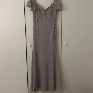  långklänning med spetsdetaljer - Elegant lila grå långklänning med v-ringad design och spetsdetaljer. Den har knappar i ryggen och sitter super bra på!! Passar perfekt till balen eller till andra fina tillfällen!! Den har aldrig kommit till användning så är i väldigt bra skick och lappen sitter fortfarande kvar!🤍🫧