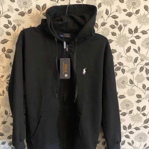 Svart hoodie  - Säljer en stilren svart hoodie från P Lauren med dragkedja och den klassiska loggan broderad i vitt på bröstet. Tröjan har en justerbar huva med snören och en praktisk känguruficka framtill. Perfekt för en avslappnad look.
