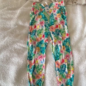 Färgglada blommiga linnebyxor - Säljer ett par färgglada byxor från H&M med ett tropiskt blommönster i grönt, rosa, blått och orange. Byxorna har en bekväm passform och är perfekta för sommaren. De har en elastisk midja för extra komfort. Inga hål. Inga fläckar. Mycket bra skick. 