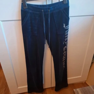 Mörkblå mjukisbyxor från Juicy Couture - Snygga mörkblå mjukisbyxor från Juicy Couture med glittrande detaljer längs benet. Byxorna har en bekväm passform med snörning i midjan och praktiska fickor både fram och bak. Perfekta för en avslappnad stil med en touch av glamour.