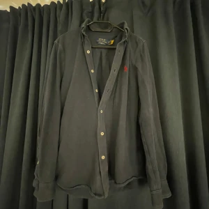 Blå skjorta från Ralph Lauren - Säljer en stilren svart skjorta från Ralph Lauren med klassisk krage och knappar framtill. Skjortan har en broderad röd logotyp på bröstet och långa ärmar. Perfekt för en elegant look. Skicket är toppen! //Svéd Resell🥇