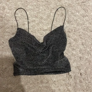 Glittrig svart linnetopp från H&M Divided - Säljer en snygg glittrig svart linnetopp från H&M Divided. Toppen har tunna axelband och vattenfalls-ringning som ger en elegant look. Perfekt för en utekväll eller festligheter. ✨