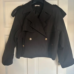 Svart trenchcoat från Musera - Snygg svart trenchcoat från Musera i en snygg design. Jackan har dubbelknäppning med knappar och en klassisk krage. Perfekt för en stilren look. Är i storlek xs men skulle säga att den sitter mer som s/m, den har små defekter med smink i kragen
