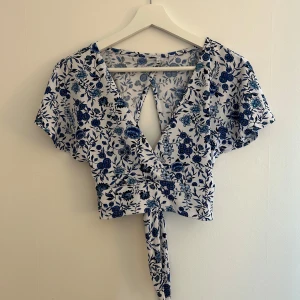 Blommig omlottopp i blått och vitt - Säljer en söt blommig omlottopp i blått och vitt. Toppen har korta ärmar och en knytning i midjan som ger en fin passform. Tryck på köp nu så postar jag så snart som möjligt! 😍