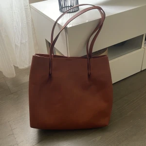 Brun axelväska i skinn - Snygg brun axelväska i skinn med enkel design och praktiska handtag. Väskan har en inre ficka med dragkedja och stängs med en tryckknapp. Perfekt för att bära med sig det nödvändigaste med stil. Perfekt till skolan/jobb😁