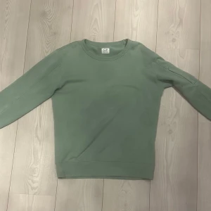 Grön tröja från C.P. Company - Säljer en stilren grön tröja från C.P. Company. Tröjan har långa ärmar och en rund halsringning. På ärmen finns en unik detalj med en rund logotyp. Perfekt för en avslappnad stil.