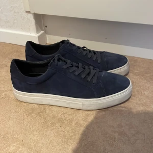 Mörkblå sneakers med vit sula - Snygga mörkblå sneakers med en stilren design och vit sula. Skorna har snörning och är tillverkade i mocka, vilket ger en lyxig känsla. Perfekta för en avslappnad men trendig look.