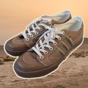 Bruna sneakers från Adidas - Snygga bruna sneakers från Adidas med klassiska ränder på sidan. Skorna har vita snören och en robust sula för extra komfort. Perfekta för en avslappnad stil.