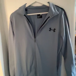 UnderArmour - Säljer en grå träningsjacka från Under Armour med dragkedja och långärmad design. Jackan har en stilren look med svart logotyp på bröstet och svarta detaljer på axlarna. Perfekt för träning eller vardagsbruk.