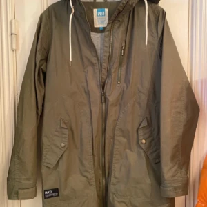 Olivgrön parkajacka från WA - Säljer en olivgrön parkajacka från WA med huva och dragkedja. Jackan har långa ärmar med justerbara muddar och flera praktiska fickor. Perfekt för höst och vår.