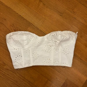 Vit axelbandslös korsettopp - Säljer en söt vit axelbandslös korsettopp med broderade blommor. Perfekt för sommardagar eller en festlig kväll. Toppen har en strukturerad passform och är både stilren och romantisk.