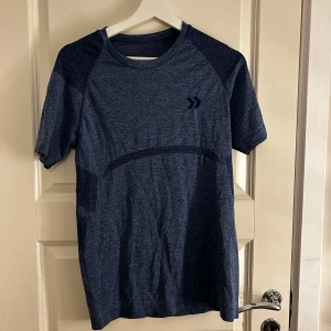 Mörkblå tränings-t-shirt - Säljer en mörkblå tränings-t-shirt med korta ärmar och rund hals. T-shirten har ett diskret mönster och är tillverkad i ett syntetmaterial som andas, perfekt för träning. Den har en liten logotyp på bröstet.