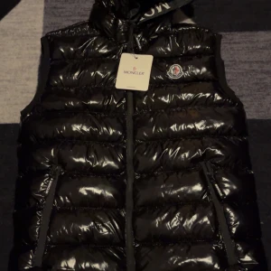 Svart dunväst från Moncler - Snygg svart dunväst från Moncler med glansig yta och dragkedja framtill. Västen har en klassisk Moncler-logga på bröstet och praktiska fickor med dragkedja. Perfekt för kyligare dagar.