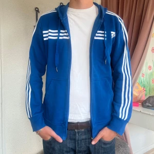 Blå huvtröja med dragkedja från Adidas - Säljer en blå huvtröja från Adidas med dragkedja och vita ränder längs ärmarna. Tröjan har en justerbar huva med snören och ribbade muddar vid ärmslut och nederkant. 