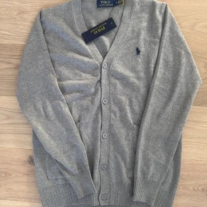 Grå kofta från Ralph Lauren - Säljer en stilren grå kofta från Ralph Lauren med knappar framtill och en broderad logotyp på bröstet. Koftan har långa ärmar och ribbade muddar vid ärmslut och nederkant. Perfekt för en klassisk look.