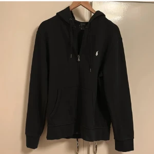 Svart hoodie från Ralph Lauren - Säljer en stilren svart hoodie från Polo Ralph Lauren med dragkedja och broderad logga på bröstet. Tröjan har långa ärmar och en praktisk huva med snörning. Perfekt för en avslappnad look.