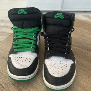 Nike Air Jordans i svart, vit och grön - Snygga Nike Air Jordans med en unik färgkombination av svart, vit och grön. Skorna har en klassisk hög design med snörning och den ikoniska Nike-loggan på sidan. Perfekta för dig som vill sticka ut med stil.