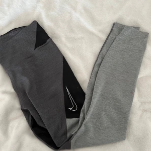 Grå och svarta leggings från Nike - Snygga grå och svarta leggings från Nike med en vit logga på benet. Perfekta för träning eller en avslappnad dag. De har en bekväm passform och är tillverkade i ett stretchigt material. En fläck finns på ena sidan, se bild