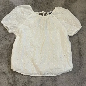 Vit blus från Vero Moda - Säljer en söt vit blus från Vero Moda i storlek S. Blusen har korta ärmar och ett fint broderat mönster. Den är tillverkad i 100% bomull och har en knytning i ryggen. Perfekt för en somrig look!