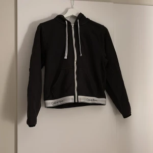 Svart huvtröja från Calvin Klein - Snygg svart huvtröja från Calvin Klein med dragkedja och vit kant med logotyp. Tröjan har långa ärmar och en praktisk huva. Perfekt för en stilren och bekväm look.