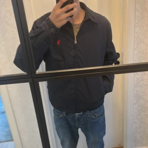 Blå jacka från U.S. Polo Assn. - Säljer en mörk blå jacka från U.S. Polo Assn. med röd logga på bröstet. Jackan har dragkedja och ett röd- och blårutigt foder. Perfekt för våren med en stilren look. Nypris 950kr