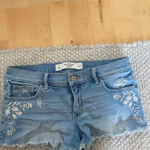 Blå jeansshorts från Abercrombie & Fitch - Snygga blå jeansshorts från Abercrombie & Fitch med fransiga kanter och broderade blommor på framsidan. Shortsen har en klassisk femficksdesign och slitna detaljer för en trendig look. Perfekta för varma sommardagar!