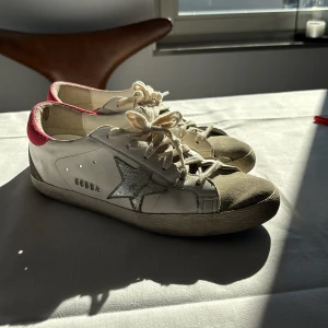 Golden Goose sneakers - Snygga Golden Goose sneakers i vitt med en grå stjärna på sidan och röd häl. Skorna har snörning och en cool, sliten look. Perfekta för en avslappnad stil.