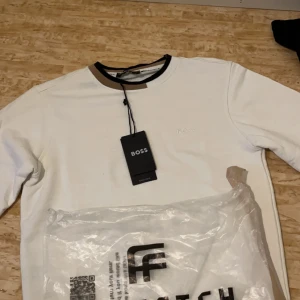 Vit tröja från BOSS - Snygg vit tröja från BOSS med subtil logga på bröstet. Tröjan har en rund halsringning med kontrasterande detaljer i svart och beige. Perfekt för en stilren look.