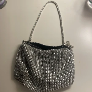 Glittrig silver axelväska - Snygg och glittrig axelväska i silver med en praktisk dragkedja. Perfekt för att lysa upp vilken outfit som helst. Väskan har en elegant design och en smidig axelrem.