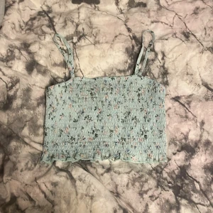 Blommig ljusblå linnetopp - Söt ljusblå linnetopp med blommigt mönster och smala axelband. Toppen har en smockad design som ger en fin passform och en lätt volangkant nedtill.