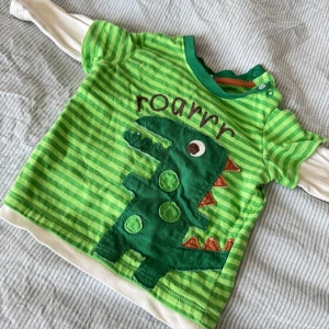 Grön randig barntröja med dinosaurie - Säljer en söt grön randig barntröja med en stor dinosaurieapplikation och texten 'roarrr' på framsidan. Tröjan har långa vita ärmar under de korta gröna och knäppning vid halsen för enkel påklädning. Perfekt för små dinosaurieälskare! 🦖Några lösa trådar förekommer enligt bil. 