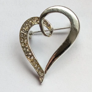 Hjärtformad brosch - Elegant hjärtformad brosch med glittrande stenar på ena sidan. Perfekt för att ge en extra touch av glamour till din outfit. Säkerhetsnål på baksidan för enkel fastsättning.