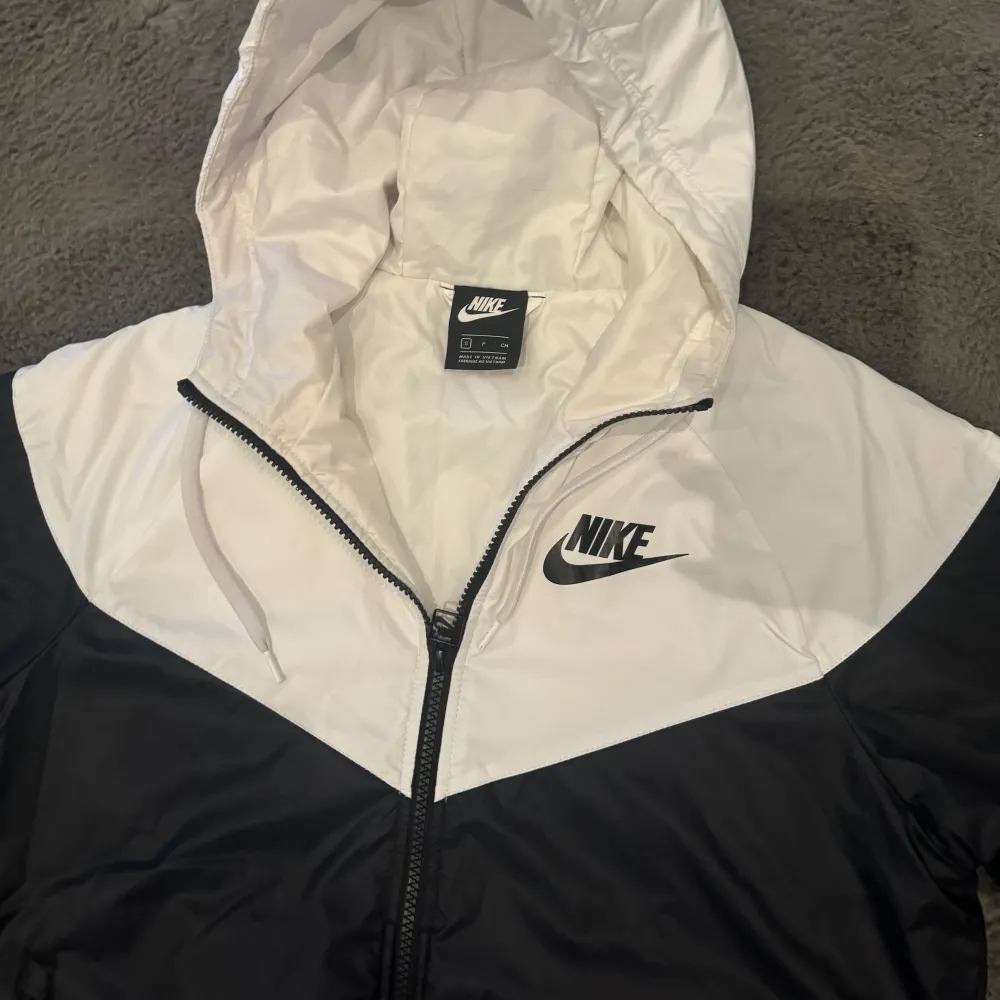 Säljer en stilren svart och vit vindjacka från Nike med huva och dragkedja. Jackan har en klassisk design med Nike-logga på bröstet och är perfekt för blåsiga dagar. Den har långa ärmar och en bekväm passform.. Takit.
