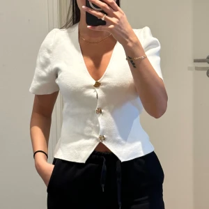 Vit kortärmad topp med knappar - Säljer en stilren vit topp med korta ärmar och dekorativa guld knappar framtill. Perfekt för en elegant och enkel look.