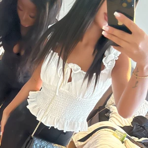 Vit ärmlös blus med volangdetaljer - Säljer en söt vit blus med volangdetaljer och knytning framtill. Blusen är ärmlös och har en smockad design som ger en fin passform. Perfekt för sommarens alla tillfällen!