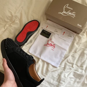 Svarta sneakers med nitar från Christian Louboutin - Snygga svarta sneakers från Christian Louboutin med ikoniska röda sulor och nitar på tån. Skorna har snörning och är tillverkade i ett elegant material som ger en lyxig känsla. Perfekta för att ge din outfit en edgy touch.
