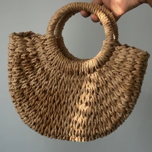 Beige semester flätad handväska - Säljer en snygg beige flätad handväska med rundade handtag. Perfekt för sommarens utflykter eller en dag på stranden. Väskan har en naturlig look och är både stilren och praktisk.