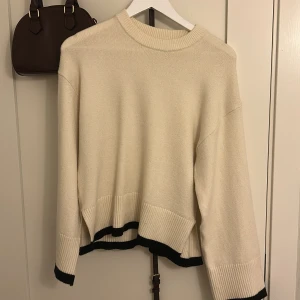 Beige stickad tröja från Gina Tricot - Säljer en stilren beige stickad tröja från Gina Tricot med svarta detaljer vid ärmslut och nederkant. Tröjan har en normal passform och långa ärmar, perfekt för kyligare dagar. Den ribbade strukturen ger en extra touch av elegans.