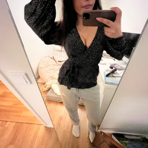 Svart omlottblus med vita prickar från & Other Stories - Säljer en snygg omlottblus med vita prickar från & Other Stories. Blusen har långa ärmar och ett knytband i midjan för en smickrande passform. Perfekt för en stilren look! Knappen på högra armen är av men enkelt ahh återgärda.