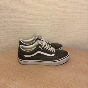 Grå sneakers från Vans - Säljer ett par klassiska grå Vans sneakers med vit sula och snörning. Skorna har den ikoniska vita sidostripen och är perfekta till våren . De är tillverkade i syntetmaterial och har en bekväm passform. Skorna är i bra skick och med en rengöring så kan man nog putsa upp dom i väldigt bra skick. Vid minsta frågor kontakta mig!
