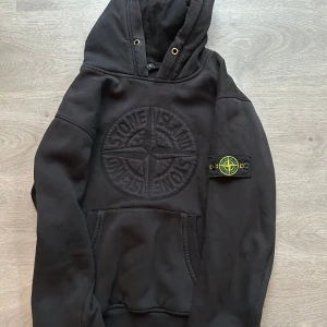 Svart hoodie från Stone Island - Säljer en svart hoodie från Stone Island med broderad logga på bröstet och ett märke på ärmen. Tröjan har en klassisk känguruficka och en justerbar huva. Perfekt för en stilren och avslappnad look.